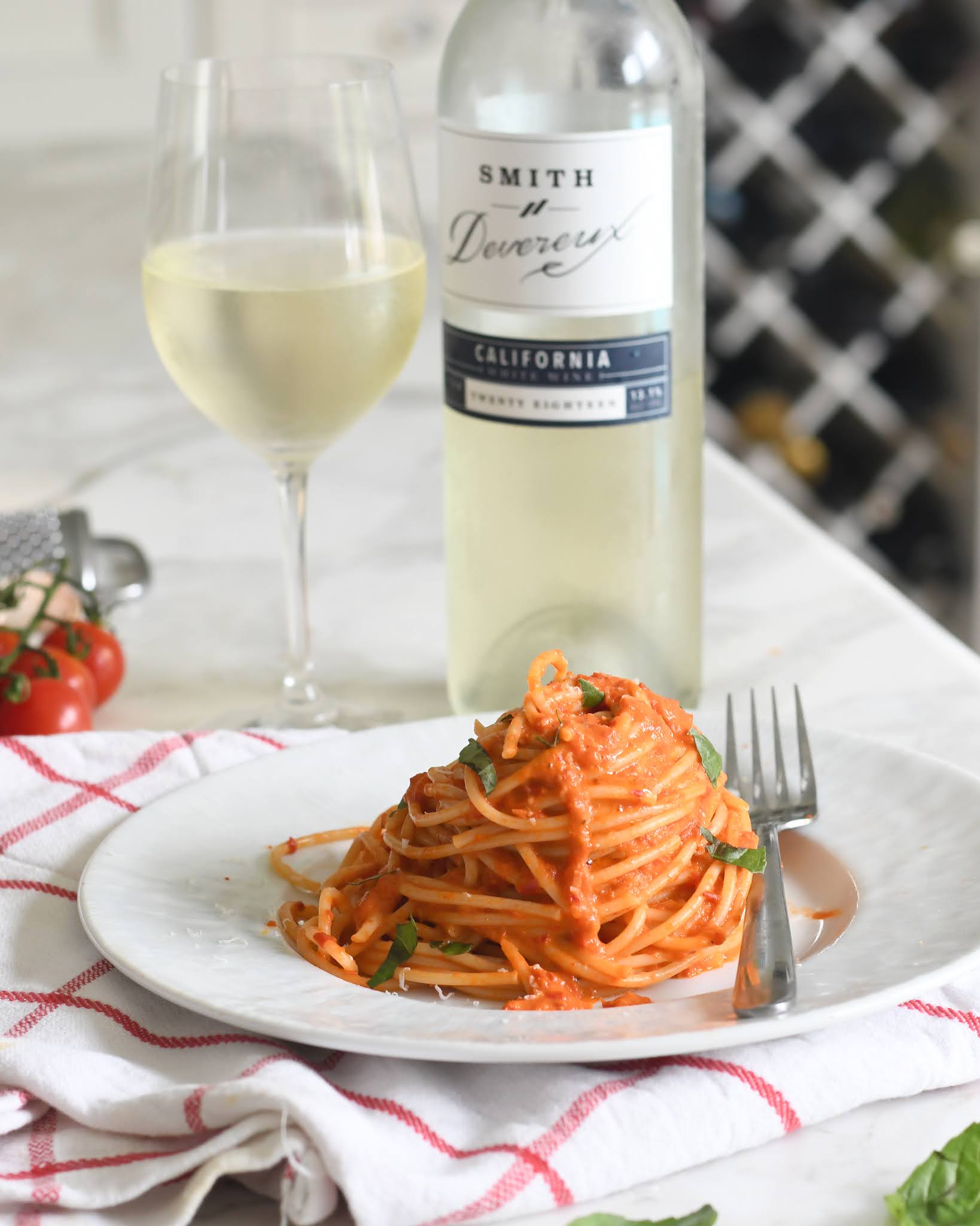 A plate of spaghetti with red sauce and herbs sits on a white plate with a fork, next to a glass and bottle of white wine on a table with a red-and-white checked cloth. Tomatoes and a cheese grater are in the background.