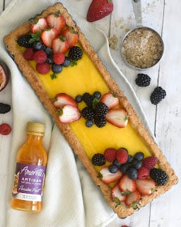A rectangular fruit tart with yellow filling is topped with strawberries, blackberries, blueberries, and mint leaves. A bottle of vanilla bean extract and a measuring cup of brown sugar are nearby on a white wooden surface.