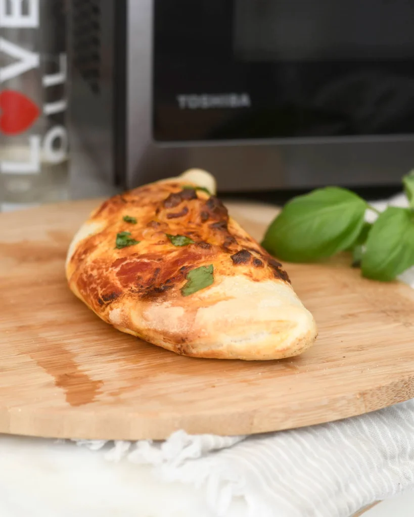 A golden-brown calzone topped with fresh basil sits on a wooden cutting board, with a basil plant and a glass of water in the background near a Toshiba microwave.