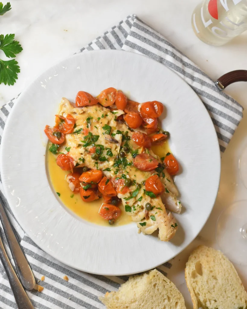 A white plate with fish fillets topped with cherry tomatoes, herbs, and sauce, placed on a striped napkin. Slices of bread, a fork and knife, a sprig of parsley, and a drink are nearby on a marble surface.