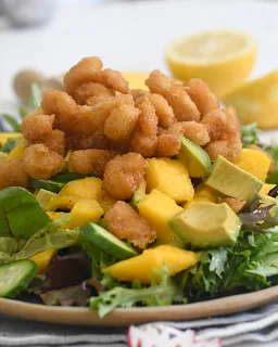 A salad topped with crispy fried shrimp, chunks of avocado, and pieces of mango, served on a bed of mixed leafy greens. A halved lemon is in the background.