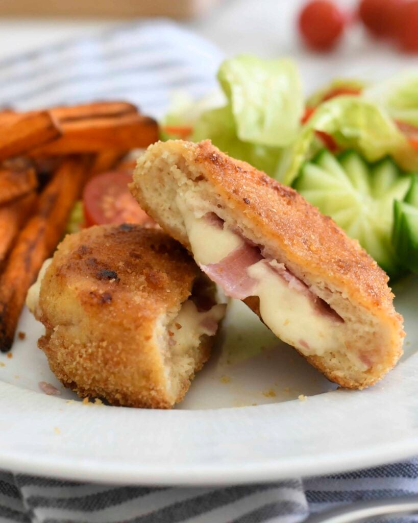 A breaded, golden-brown chicken cordon bleu is sliced in half, revealing melted cheese and ham inside. Sweet potato fries and a fresh salad with cucumber, lettuce, and tomato are on the side.