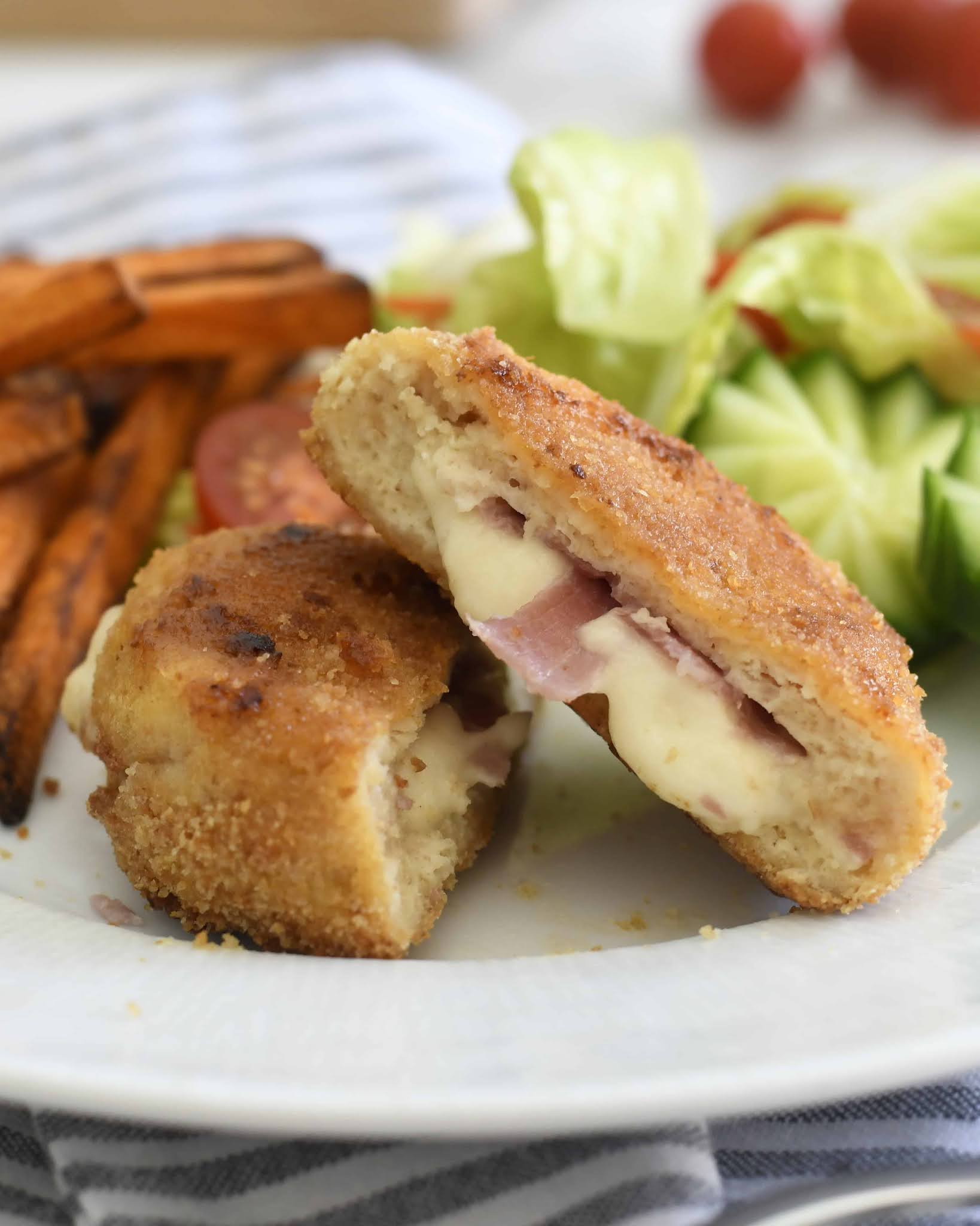 A breaded, golden-brown chicken cordon bleu is sliced in half, revealing melted cheese and ham inside. Sweet potato fries and a fresh salad with cucumber, lettuce, and tomato are on the side.