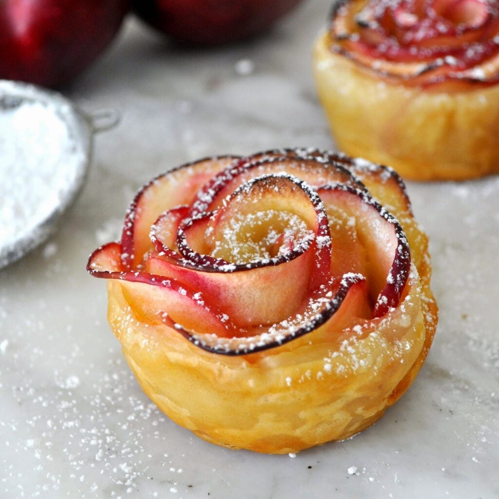 A pastry shaped like a rose made from thin apple slices, dusted with powdered sugar, sits on a marble surface with another similar pastry and apples in the background.