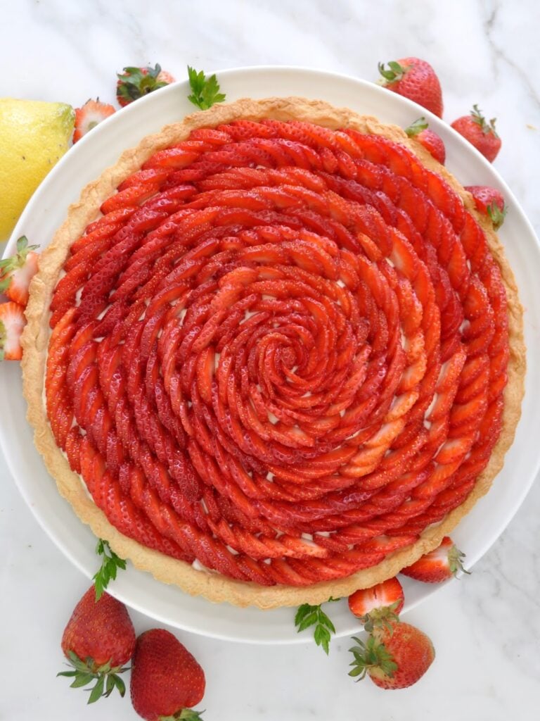 A strawberry tart with thinly sliced strawberries arranged in a spiral pattern on top of a golden crust, surrounded by fresh strawberries on a white plate and marble surface.