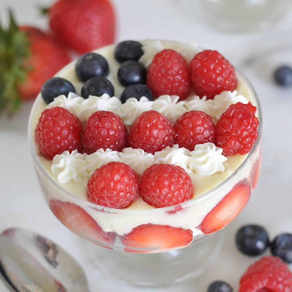 A glass dessert dish layered with cream, sliced strawberries, and topped with rows of fresh raspberries, blueberries, and piped whipped cream. Whole strawberries and blueberries are scattered around the dish.