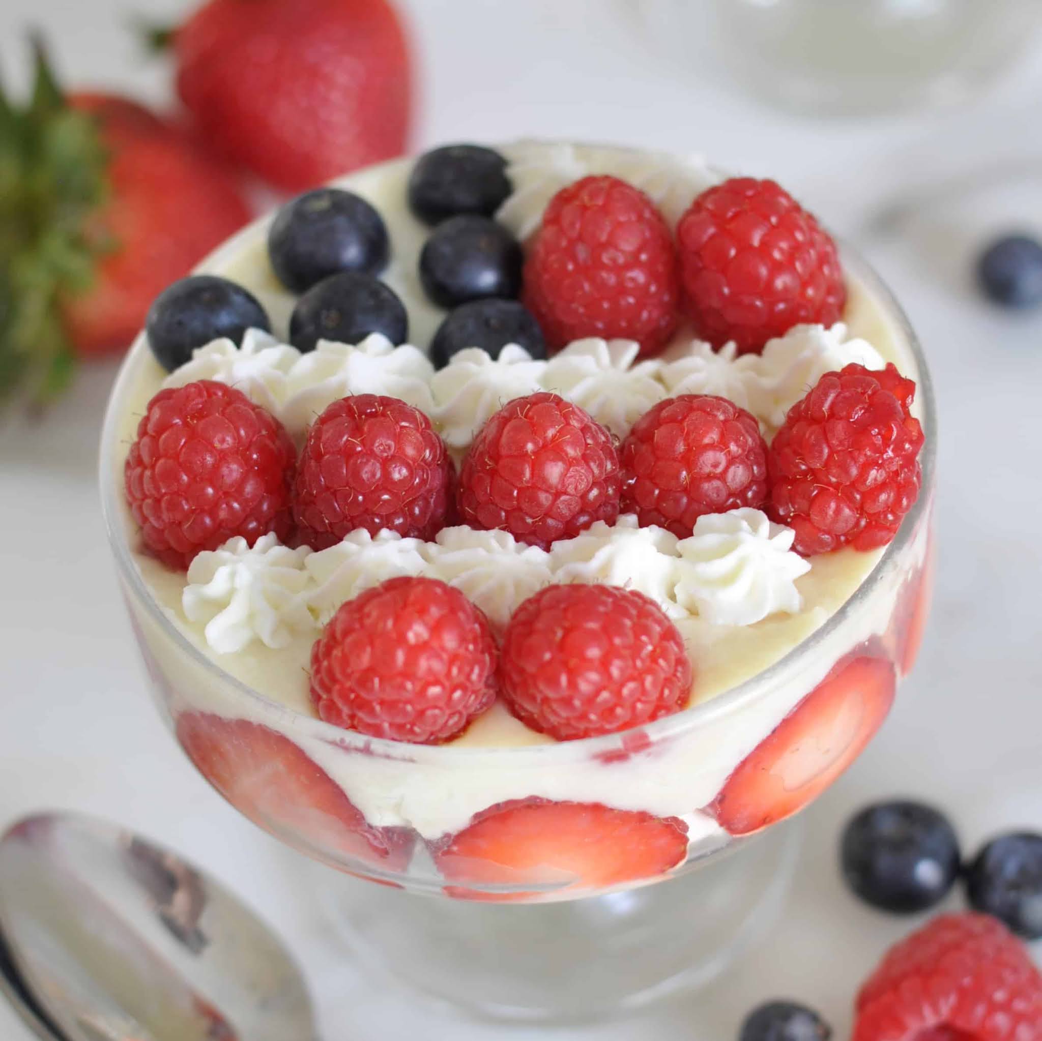 A glass dessert dish layered with cream, sliced strawberries, and topped with rows of fresh raspberries, blueberries, and piped whipped cream. Whole strawberries and blueberries are scattered around the dish.