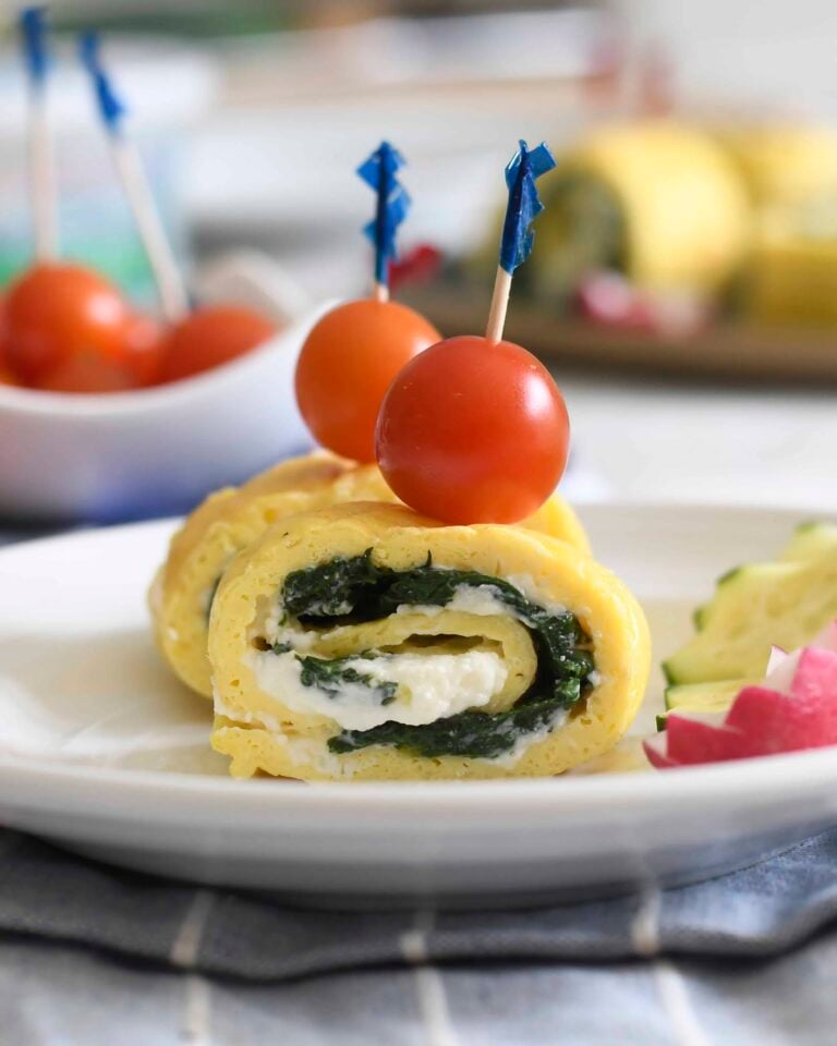 A rolled omelet slice filled with spinach and cheese is garnished with a skewer holding two cherry tomatoes, presented on a white plate with avocado and radish slices on the side.