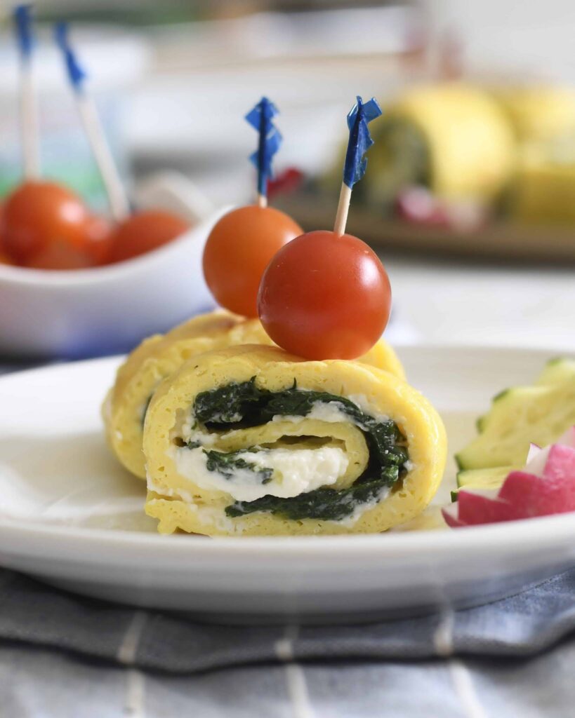 A rolled omelet slice filled with spinach and cheese is garnished with a skewer holding two cherry tomatoes, presented on a white plate with avocado and radish slices on the side.