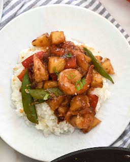 A plate of white rice topped with shrimp, pineapple chunks, snap peas, and red bell peppers in a brown sauce, garnished with chopped green onions.