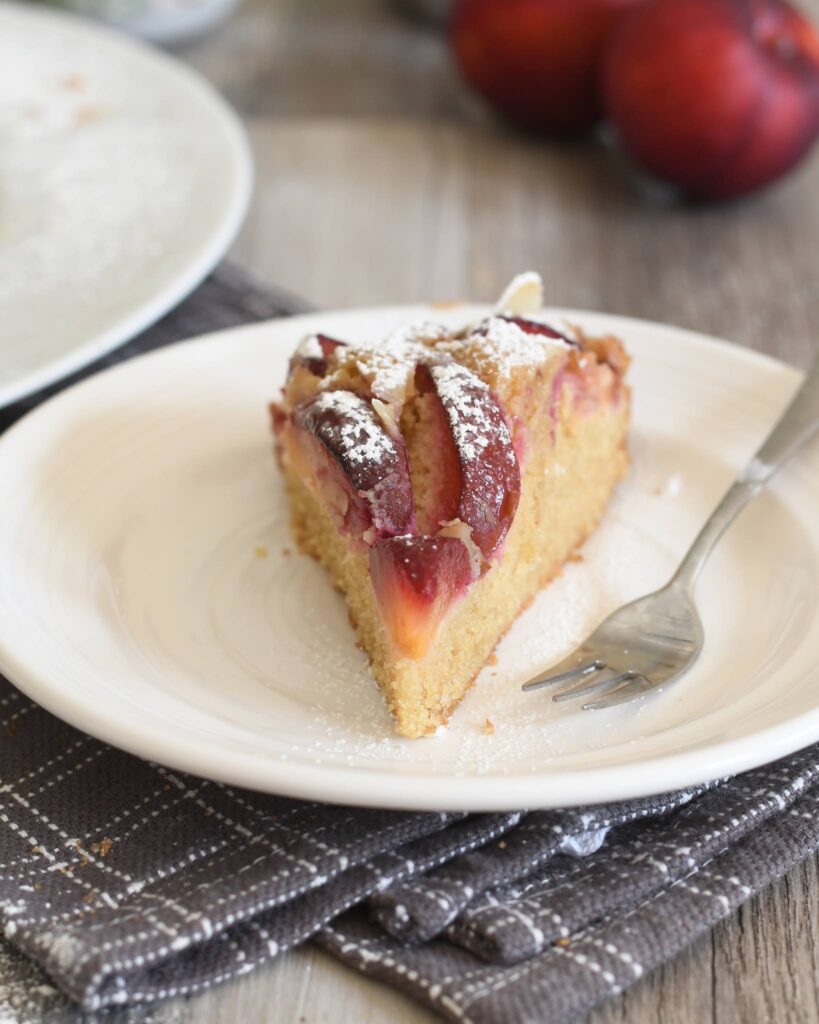 A slice of plum cake topped with powdered sugar sits on a white plate with a fork, placed on a brown checkered napkin. Whole plums and another plate are blurred in the background.