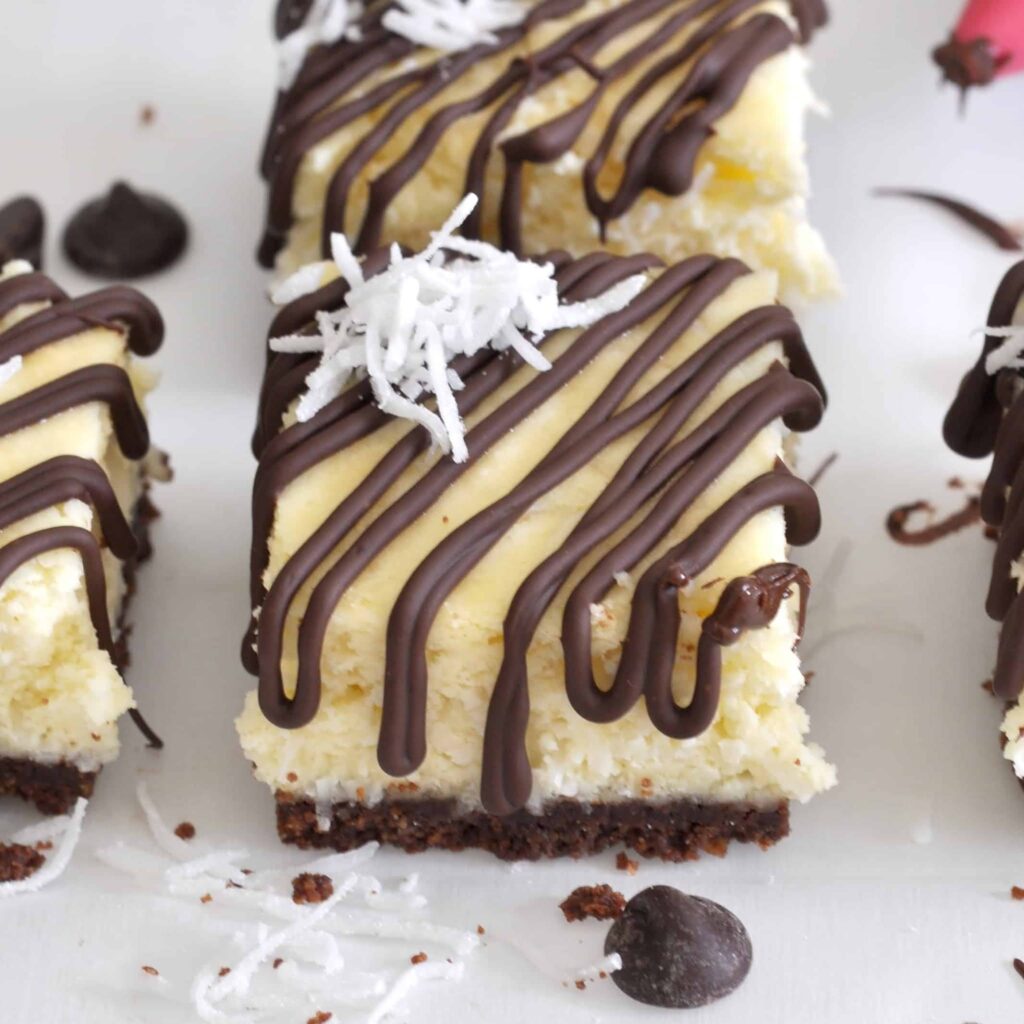 A close-up of coconut cheesecake bars with a chocolate crust, topped with drizzled dark chocolate and shredded coconut, arranged on a white surface with scattered chocolate chips and coconut flakes.