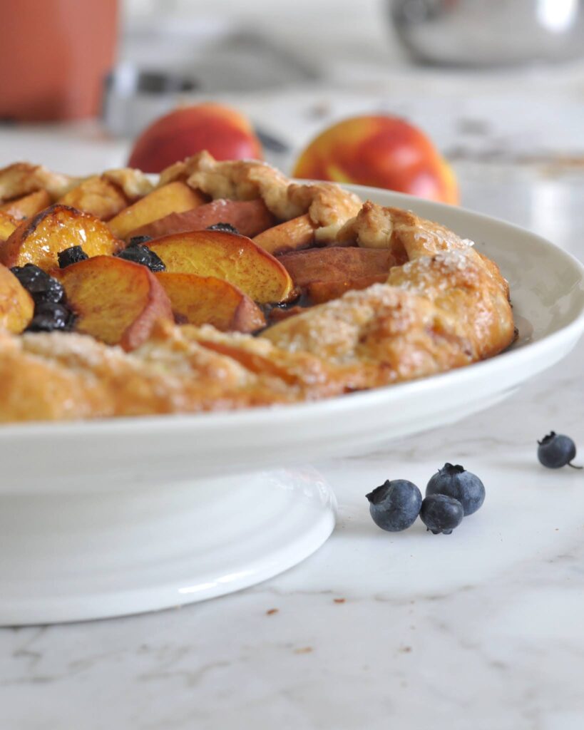 A rustic peach and blueberry galette sits on a white cake stand, with fresh peaches and blueberries scattered on a marble countertop in the background.