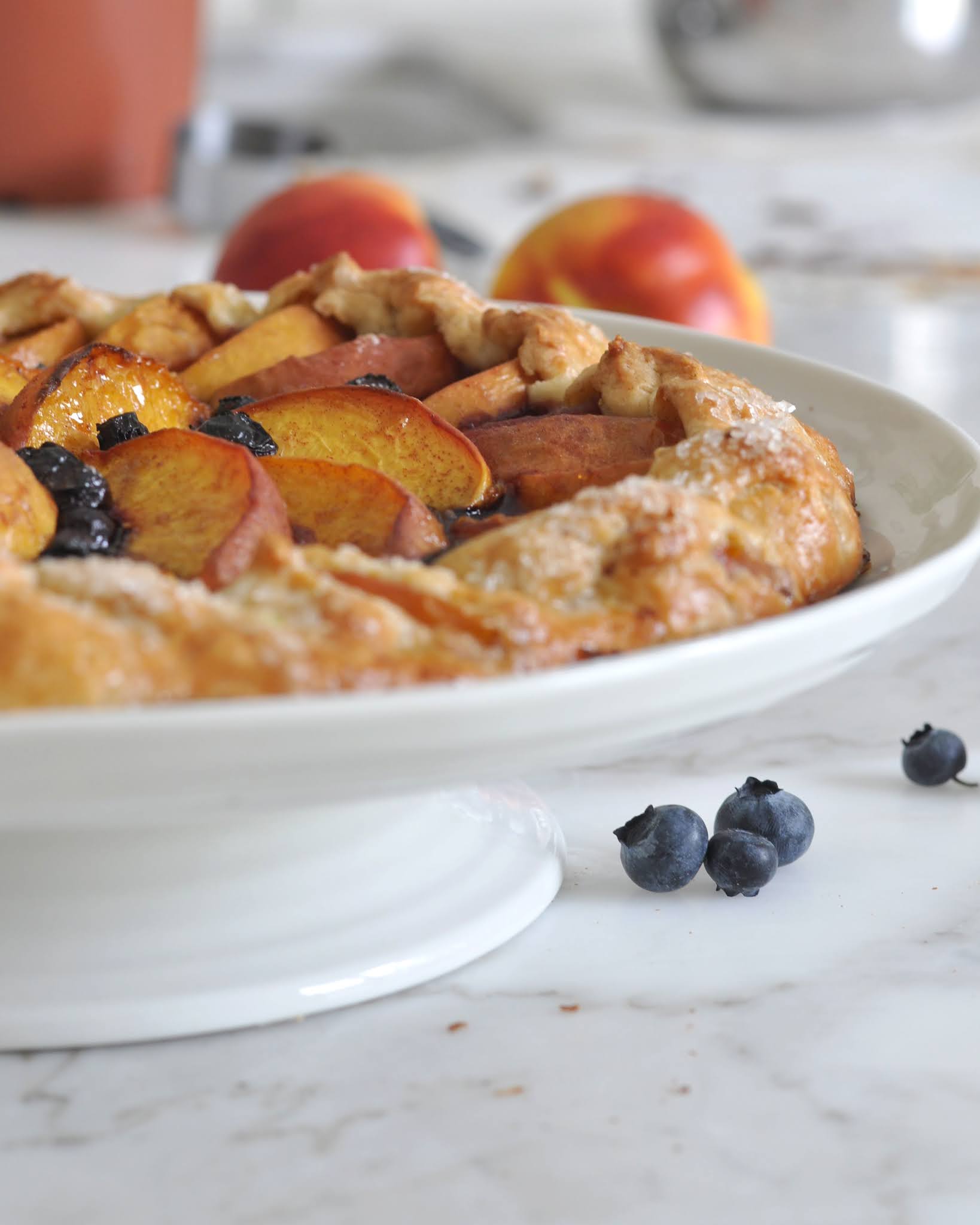 A rustic peach and blueberry galette sits on a white cake stand, with fresh peaches and blueberries scattered on a marble countertop in the background.