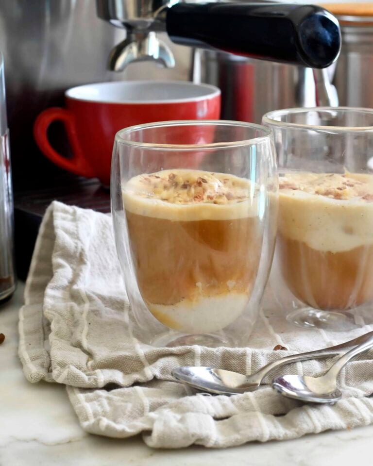 Two clear double-walled glasses with affogato—espresso poured over vanilla ice cream—sit on a folded cloth next to spoons, with a red cup and espresso machine in the background.