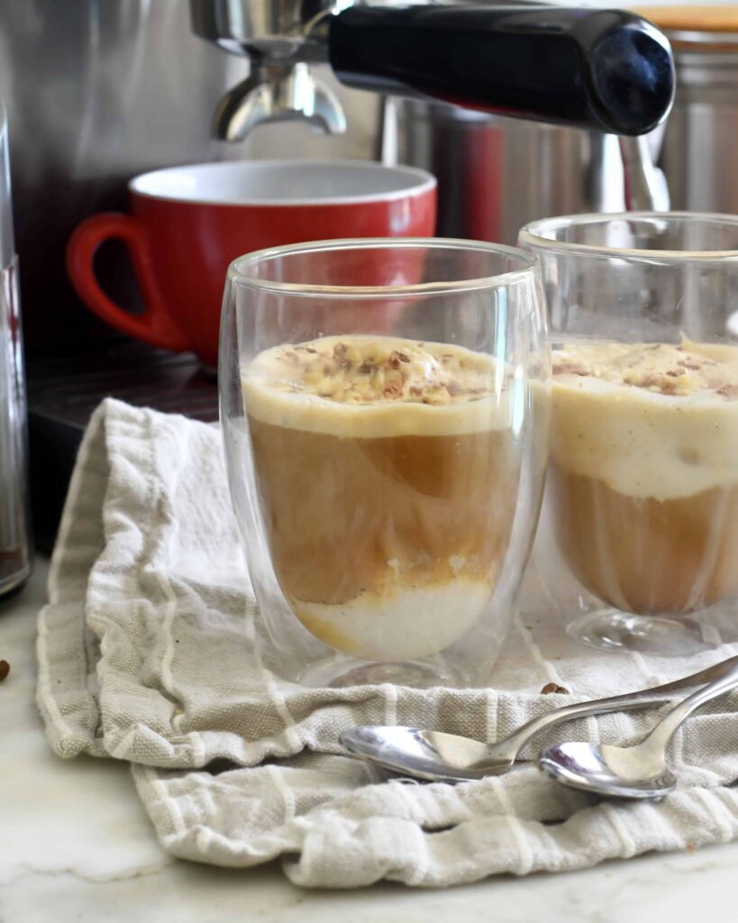 Two clear double-walled glasses with affogato—espresso poured over vanilla ice cream—sit on a folded cloth next to spoons, with a red cup and espresso machine in the background.