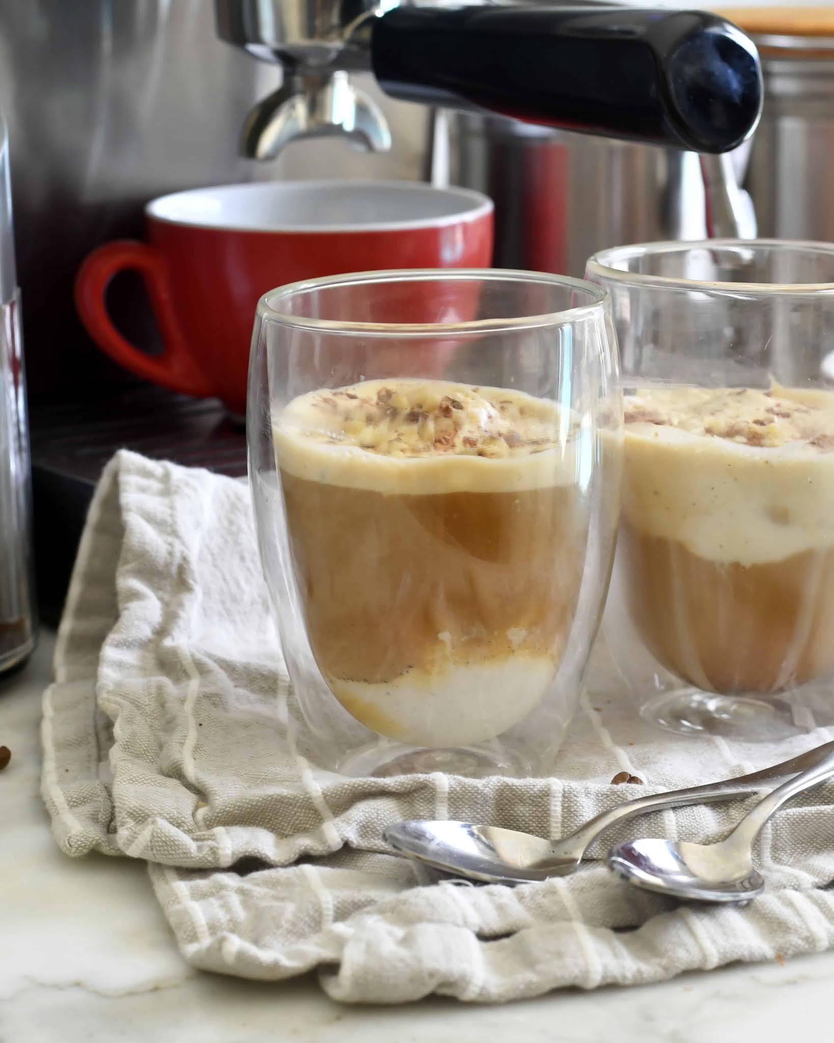 Two clear double-walled glasses with affogato—espresso poured over vanilla ice cream—sit on a folded cloth next to spoons, with a red cup and espresso machine in the background.