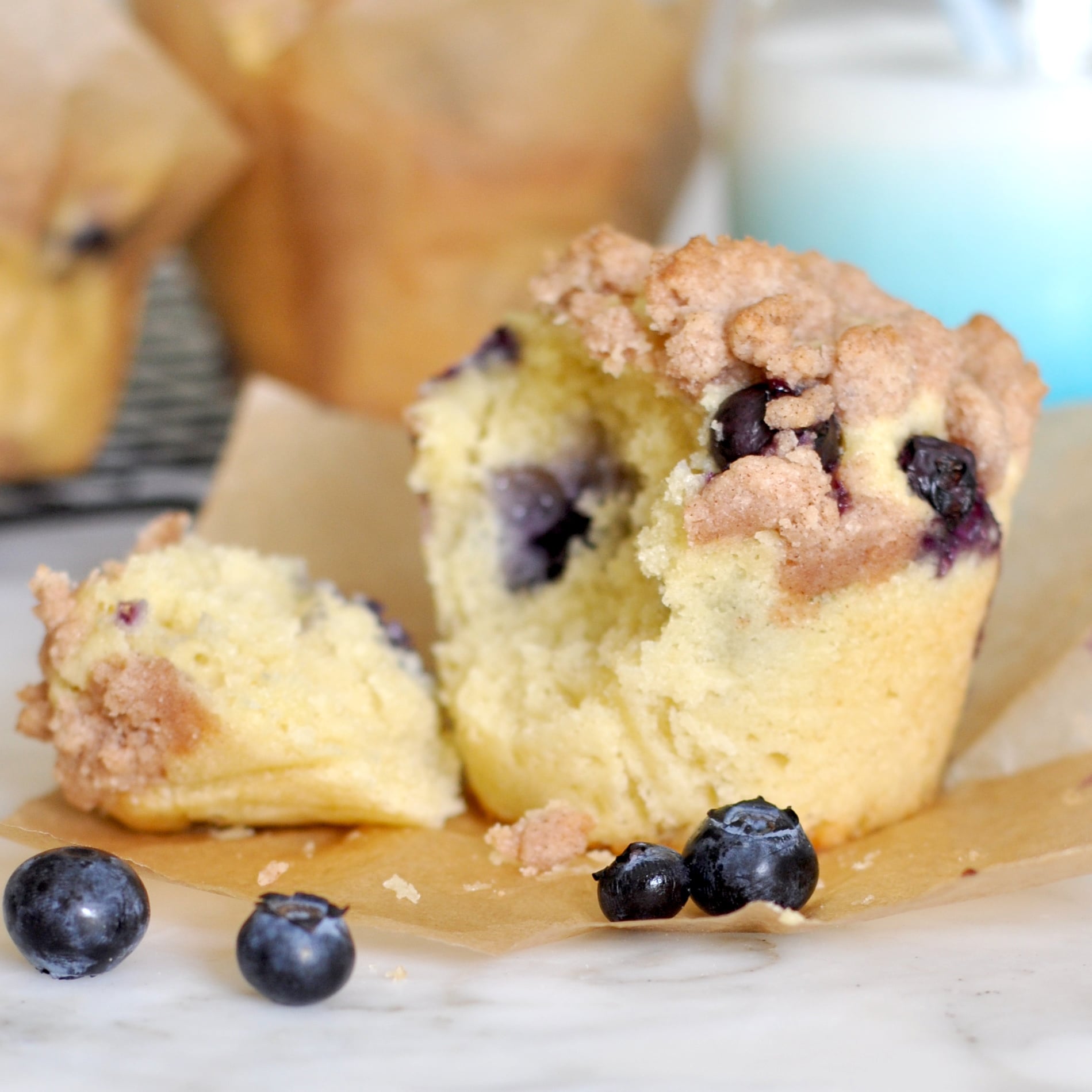 A blueberry muffin with a crumb topping is broken in half on parchment paper, with fresh blueberries scattered nearby. A glass of milk is blurred in the background.