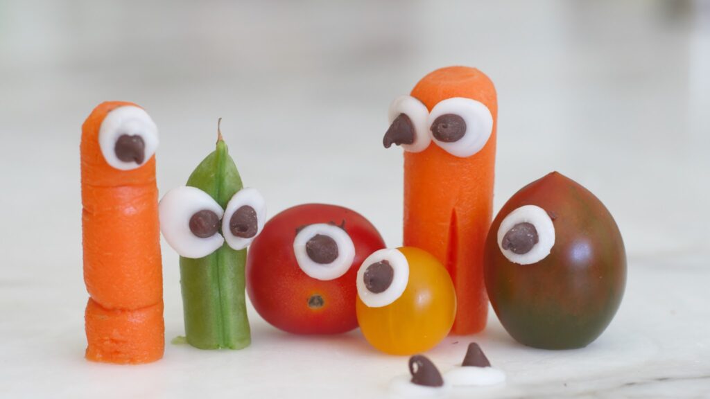 Vegetables and cherry tomatoes with candy eyes arranged to resemble cartoonish, surprised faces on a white surface.