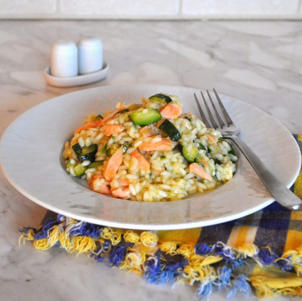 A plate of risotto with pieces of zucchini and salmon, served with a fork on the side. The plate sits on a colorful fringed napkin on a marble countertop, with salt and pepper shakers in the background.