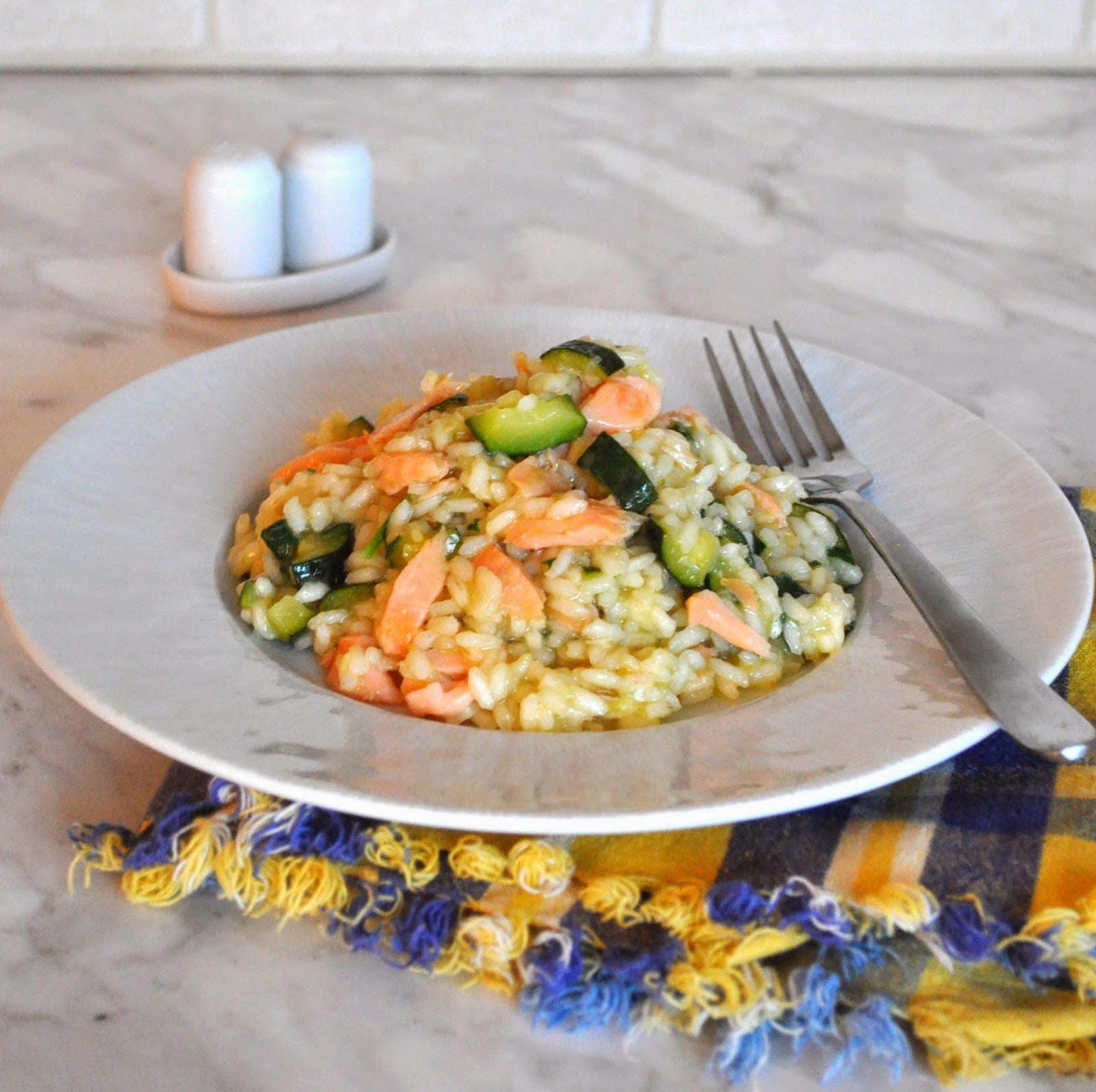A plate of risotto with pieces of zucchini and salmon, served with a fork on the side. The plate sits on a colorful fringed napkin on a marble countertop, with salt and pepper shakers in the background.