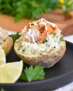 A baked potato half filled with chopped herbs, creamy topping, and crispy bits, served on a black plate with lime wedges and parsley garnish.