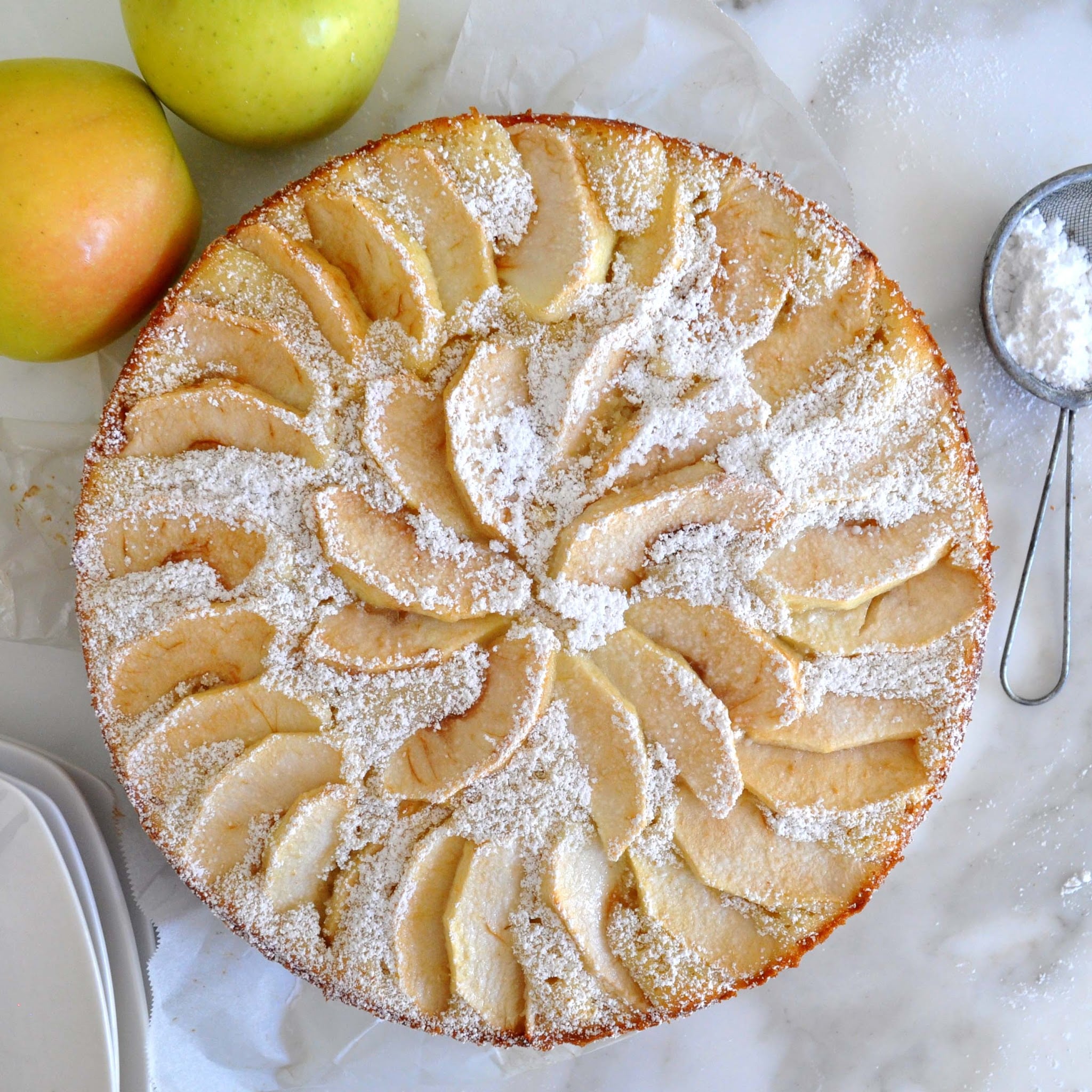 Top-down view of a round apple cake topped with sliced apples and dusted with powdered sugar, placed on parchment paper next to two apples and a sifter with powdered sugar on a marble surface.