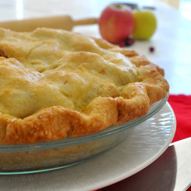 A freshly baked pie with a golden, flaky crust sits in a glass pie dish on a white plate. In the background, there are apples and a rolling pin on a light-colored surface.