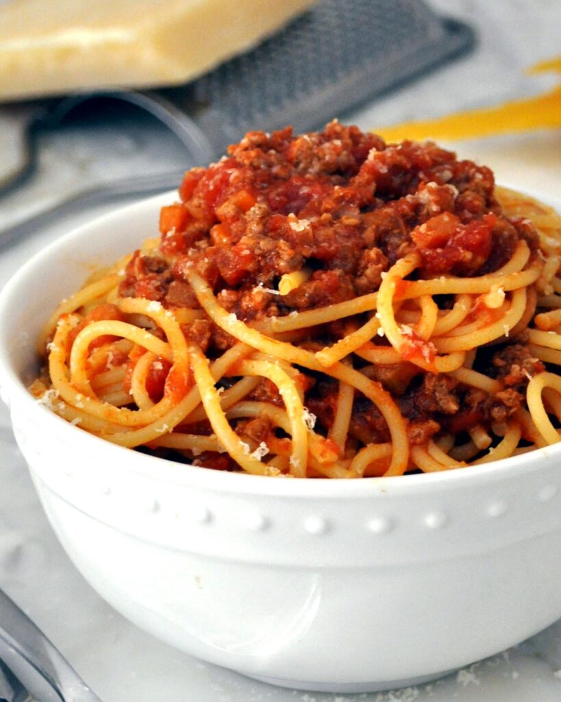 A white bowl filled with spaghetti topped with a rich, chunky tomato and ground meat sauce, sits on a marble surface. In the background, a block of cheese and a grater are visible.