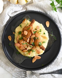 A black plate with mashed potatoes topped with two pieces of chicken, garnished with toasted almond slices and chopped parsley, set on a light cloth with a fork nearby.