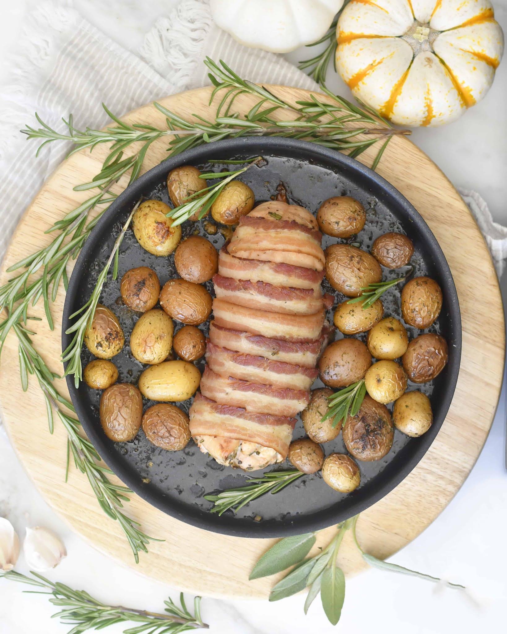 A round black pan with bacon-wrapped meatloaf and roasted baby potatoes, garnished with fresh rosemary. The pan sits on a wooden board with a small pumpkin and herbs nearby.