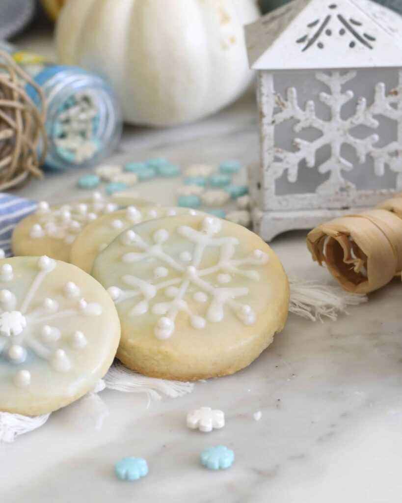 Round sugar cookies decorated with white icing snowflakes are displayed on a marble surface, surrounded by blue and white sprinkles, a small white lantern with a snowflake design, and rustic holiday decorations.