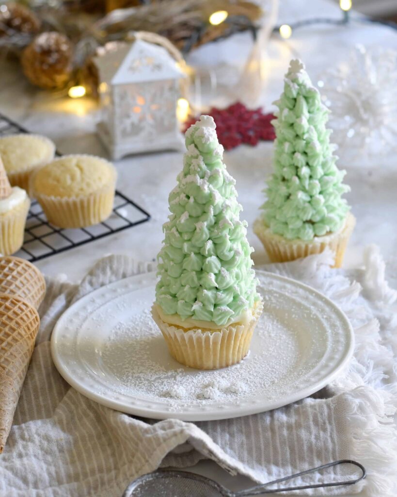 A cupcake decorated with green frosting shaped like a Christmas tree sits on a white plate, dusted with powdered sugar, with more decorated cupcakes and cones in the background on a festive table.