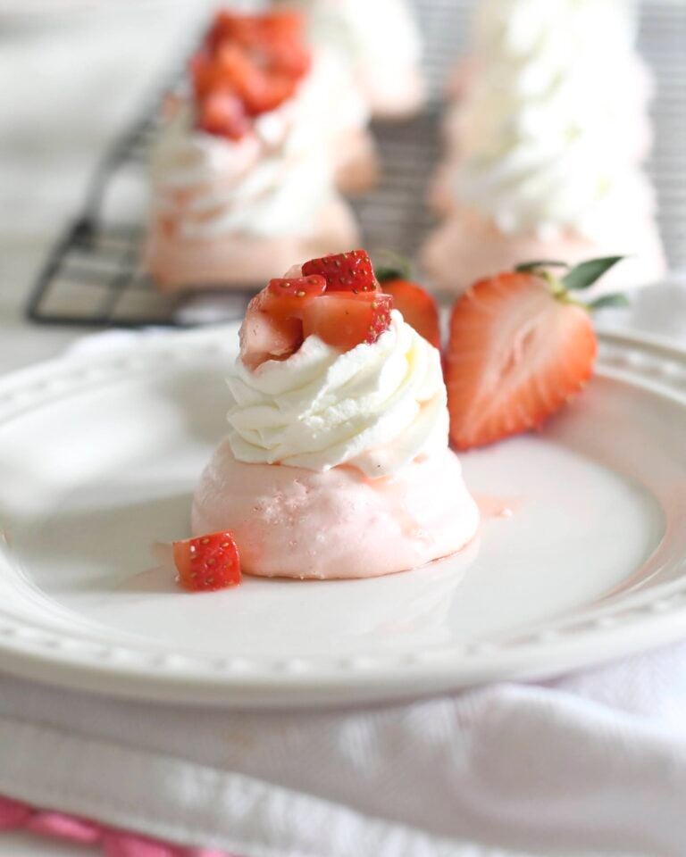 A small pink meringue topped with whipped cream and diced strawberries sits on a white plate, garnished with a halved strawberry. More similar desserts are visible on a cooling rack in the background.