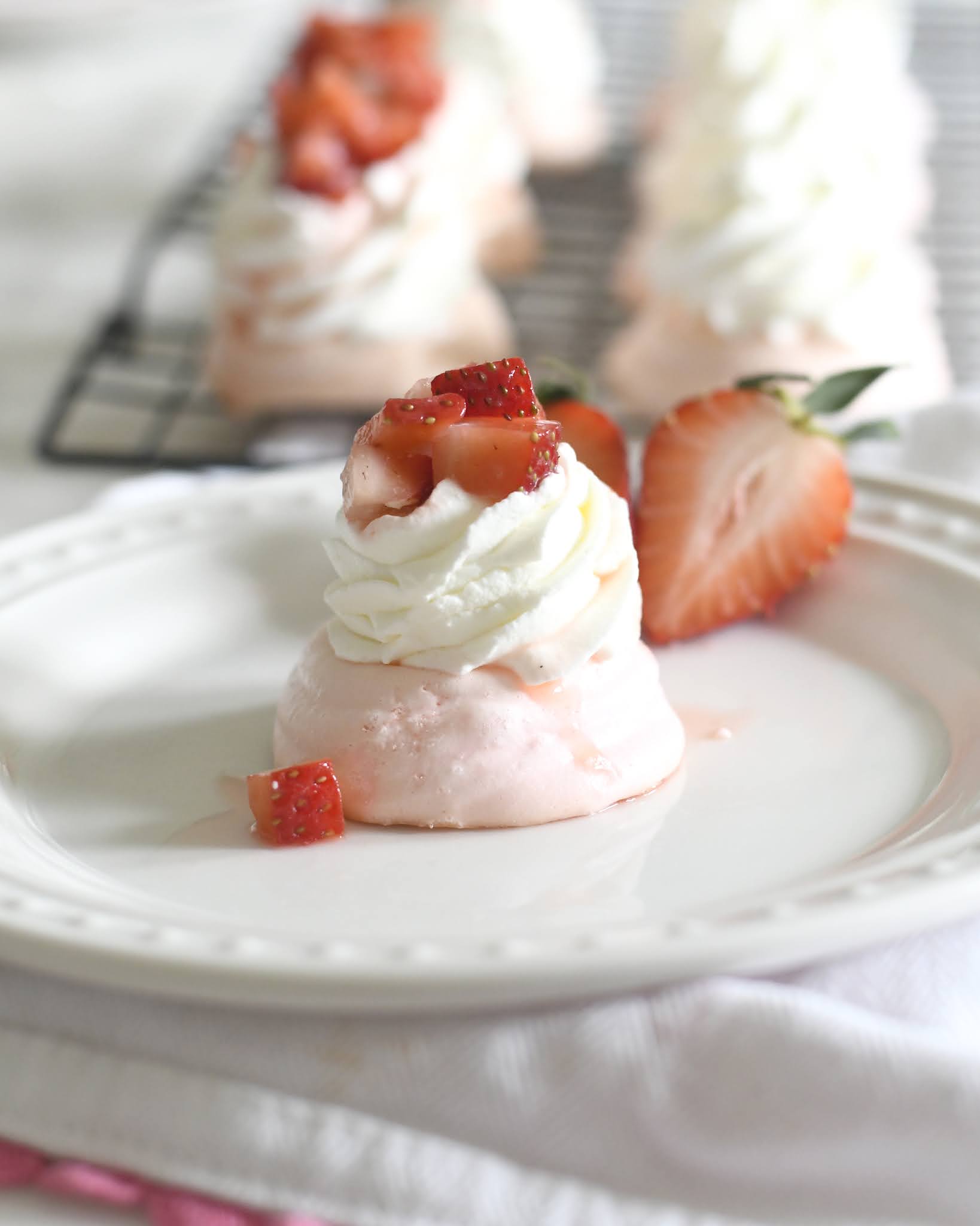 A small pink meringue topped with whipped cream and diced strawberries sits on a white plate, garnished with a halved strawberry. More similar desserts are visible on a cooling rack in the background.