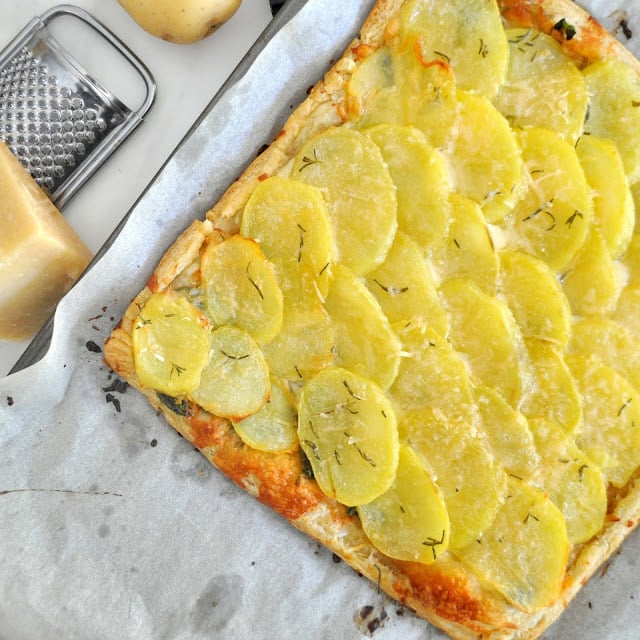 A rectangular potato tart on parchment paper, topped with thinly sliced yellow potatoes and herbs. A cheese grater, a block of cheese, and a whole potato are visible nearby on the countertop.