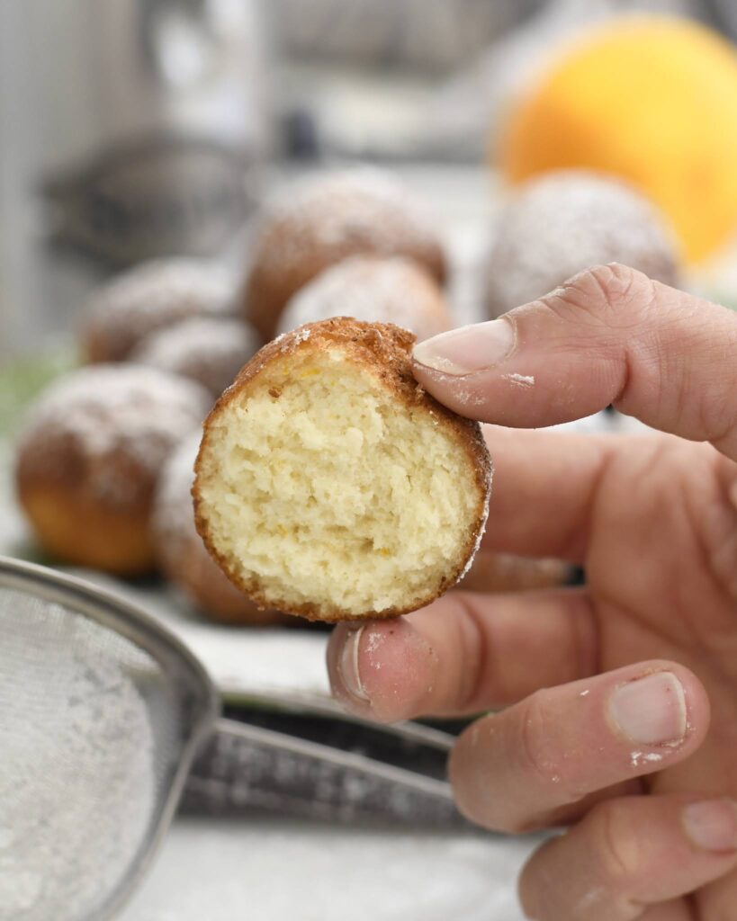 A hand holds a small, round donut with a bite taken out, showing its fluffy inside. Other powdered donuts and a metal sifter are blurred in the background.
