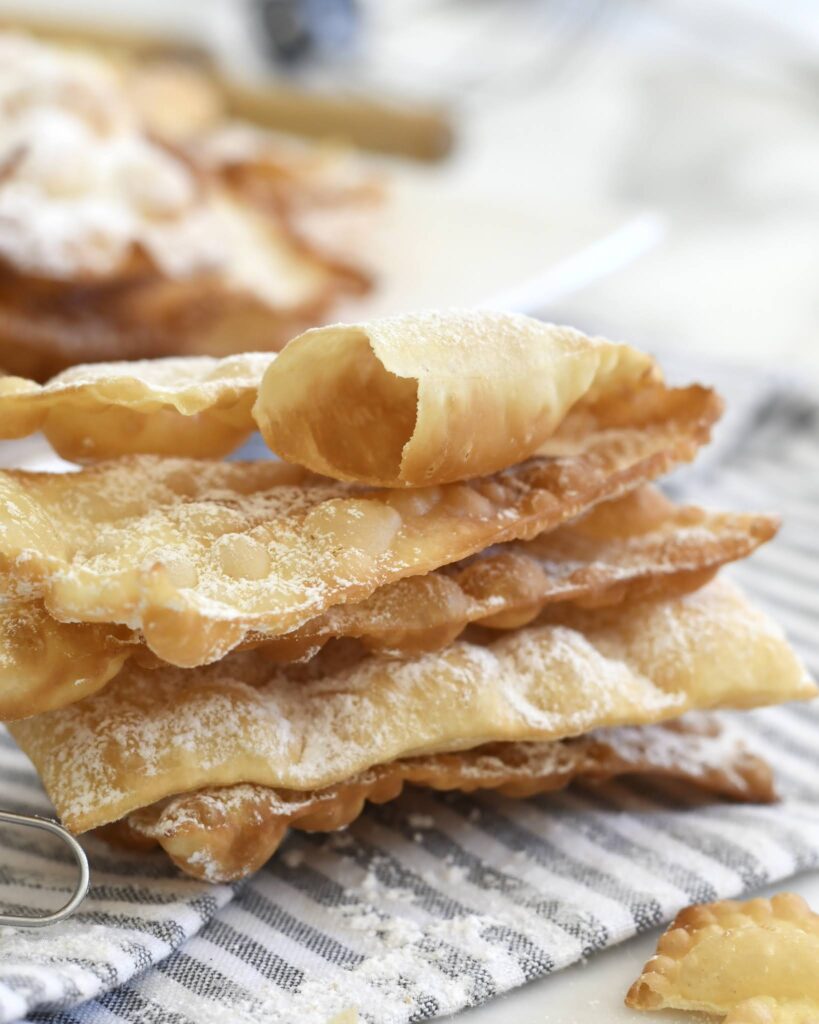 A stack of thin, crispy Italian pastry pieces dusted with powdered sugar sits on a striped cloth, with more pastries blurred in the background.
