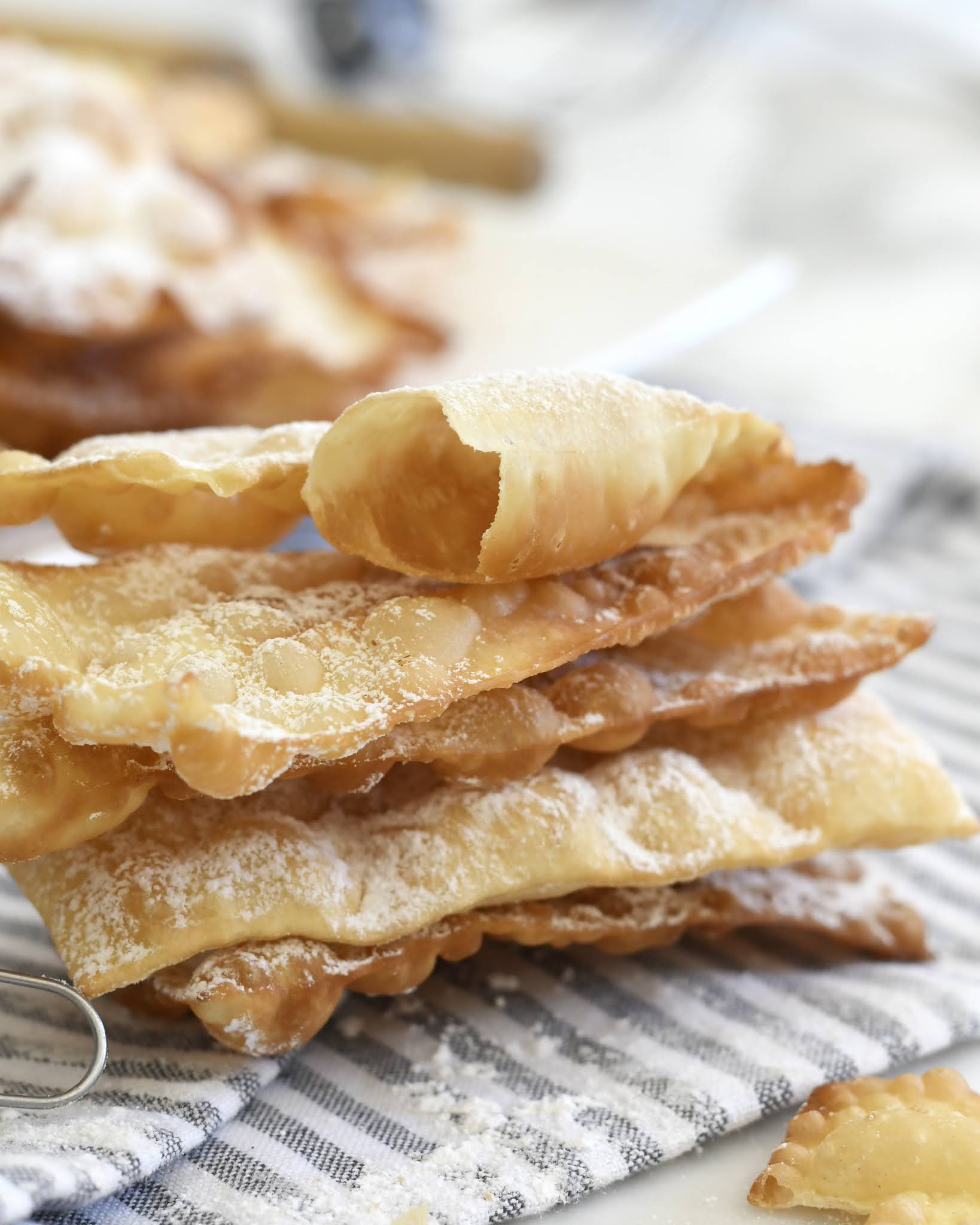 A stack of thin, crispy Italian pastry pieces dusted with powdered sugar sits on a striped cloth, with more pastries blurred in the background.