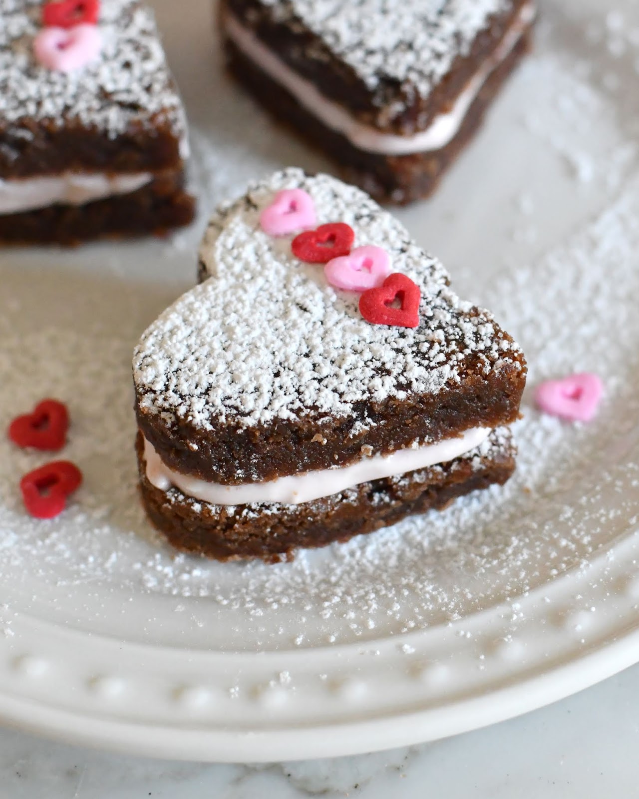 A heart-shaped brownie sandwich with powdered sugar on top, filled with cream, and decorated with small pink and red heart sprinkles, sits on a white plate.