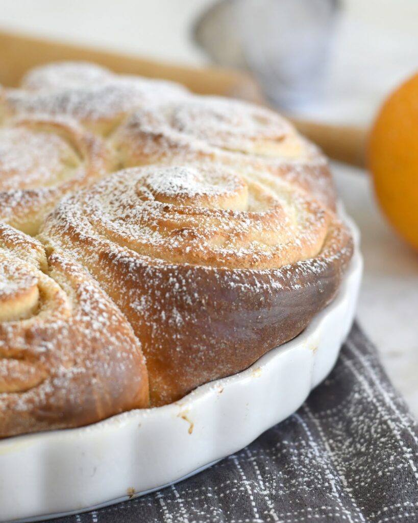 A close-up of golden brown, freshly baked cinnamon rolls in a white scalloped dish, dusted with powdered sugar, resting on a gray checkered cloth. An orange and rolling pin are blurred in the background.
