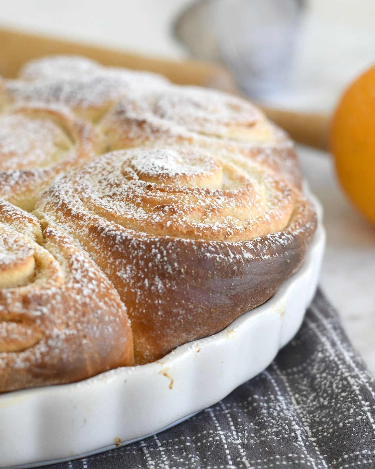 A close-up of golden brown, freshly baked cinnamon rolls in a white scalloped dish, dusted with powdered sugar, resting on a gray checkered cloth. An orange and rolling pin are blurred in the background.