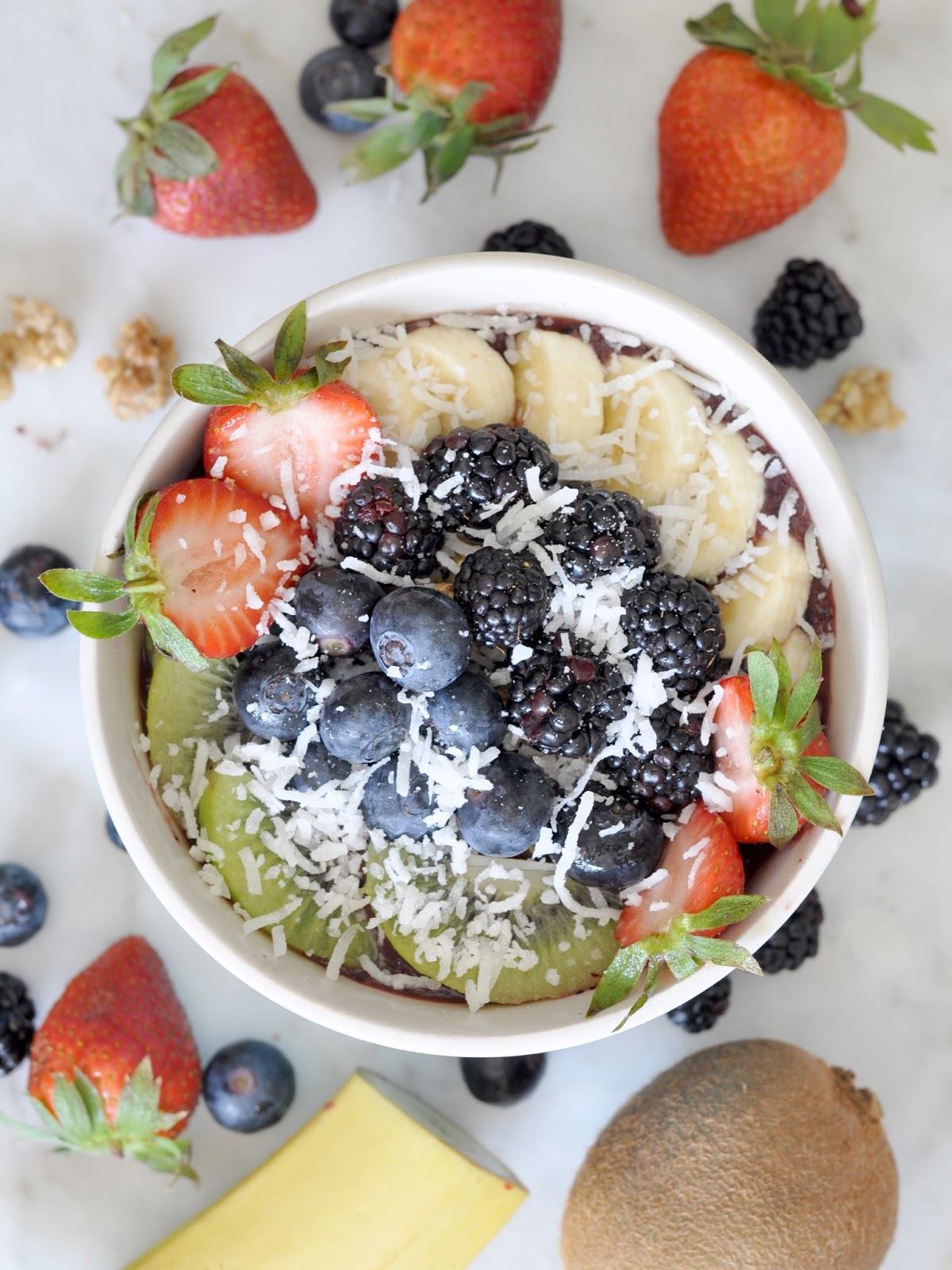 A smoothie bowl topped with sliced bananas, strawberries, blueberries, blackberries, kiwi, and shredded coconut. Surrounding the bowl are more strawberries, blueberries, blackberries, a kiwi, and part of a banana.
