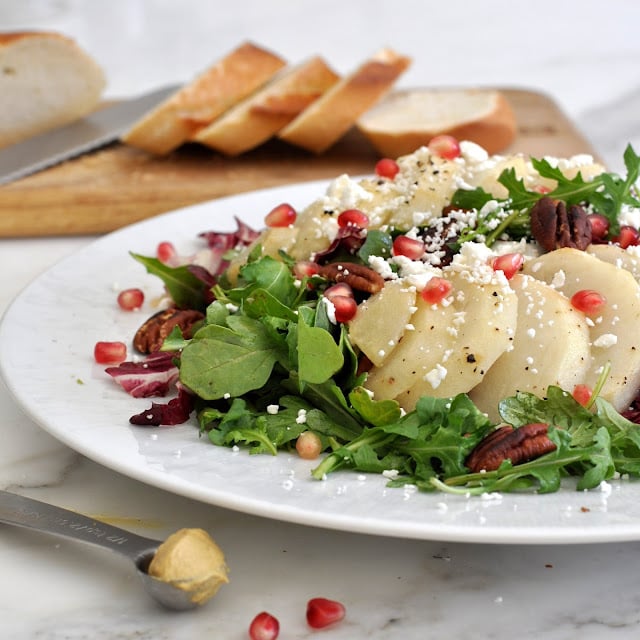 A salad with sliced pears, arugula, pecans, pomegranate seeds, and crumbled cheese on a white plate. In the background, sliced bread and a knife are on a wooden board. A spoon with mustard is in the foreground.