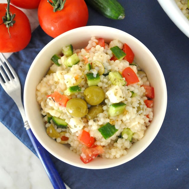 A bowl of couscous salad with diced tomatoes, cucumbers, feta cheese, and green olives, placed on a blue napkin beside whole tomatoes and a fork.