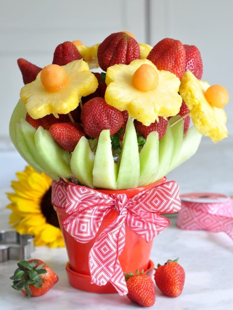 A fruit bouquet in a red pot, featuring strawberries, pineapple flowers with melon centers, and honeydew slices, all tied with a red and white geometric ribbon. A sunflower and ribbon are in the background.