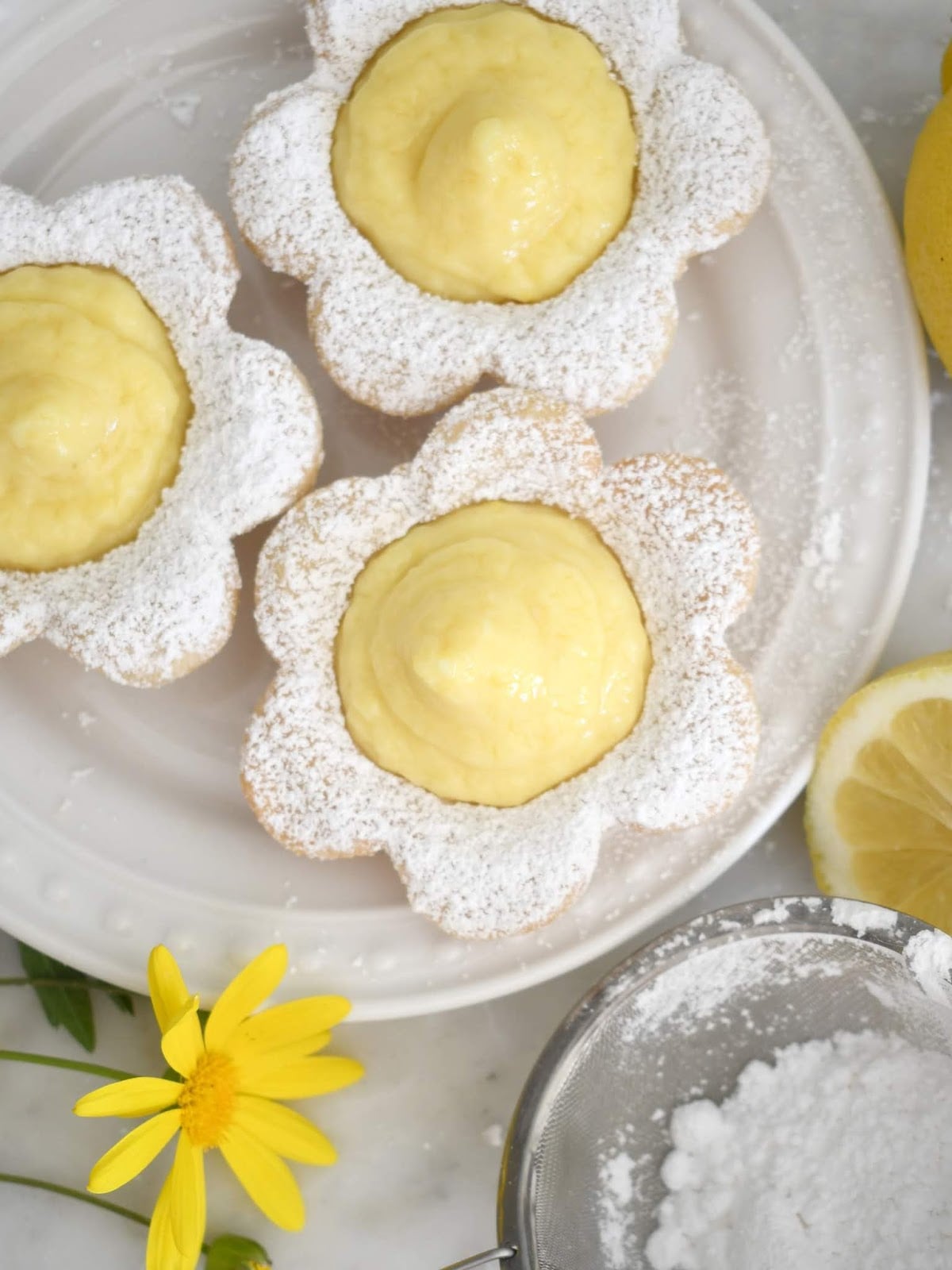 Three flower-shaped cookies filled with lemon cream and dusted with powdered sugar sit on a white plate. A yellow flower, a lemon slice, and a sifter with powdered sugar are nearby on a marble surface.