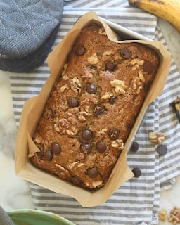 A loaf of banana bread with chocolate chips and walnuts sits in a parchment-lined pan on a striped cloth, with a banana, scattered walnuts, and chocolate chips nearby.