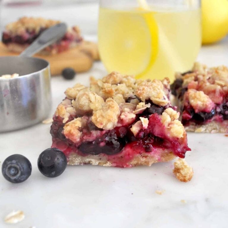 A close-up of a berry crumble bar with oats, surrounded by fresh blueberries, a metal measuring cup, and a glass of lemonade with lemon slices in the background. The bar has a crumbly topping and vibrant berry filling.