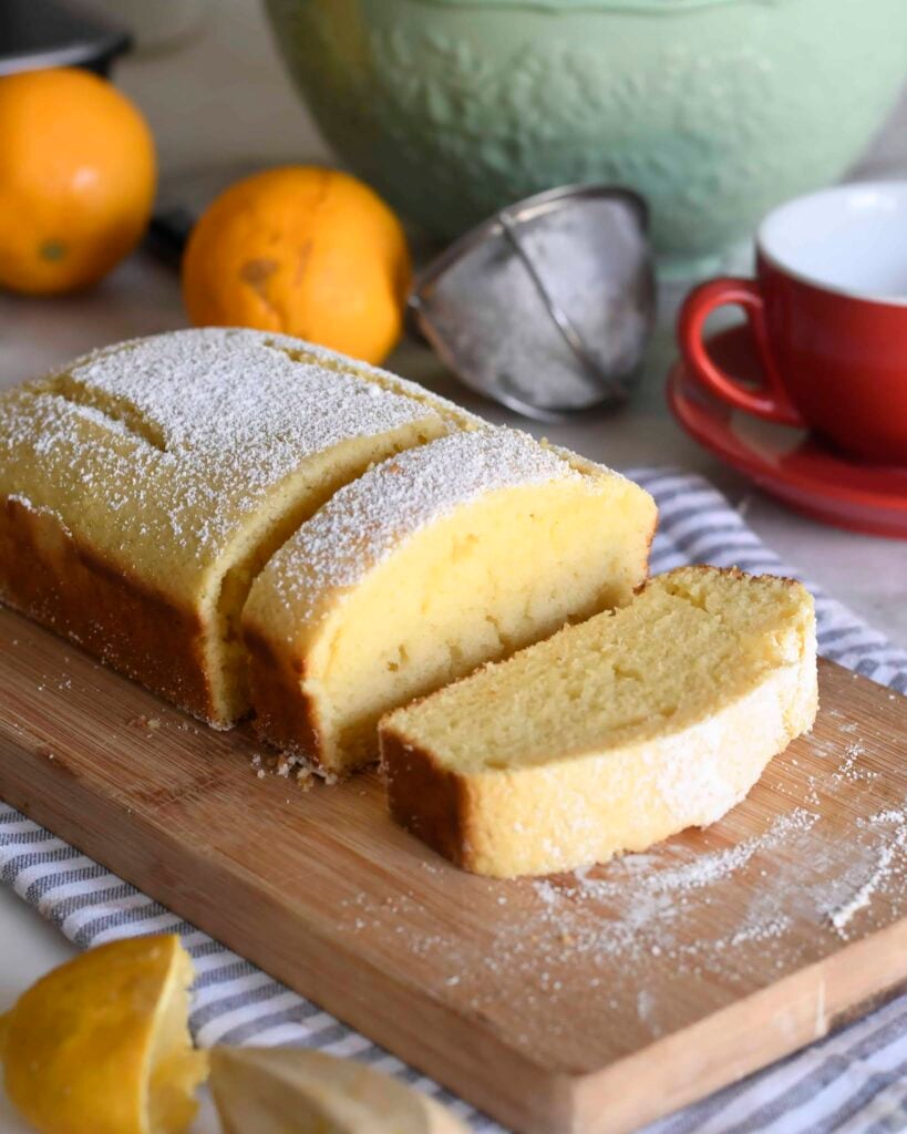A loaf of lemon cake dusted with powdered sugar sits on a wooden cutting board, with one slice cut. Nearby are fresh lemons, a green mixing bowl, a red cup and saucer, and a citrus juicer.
